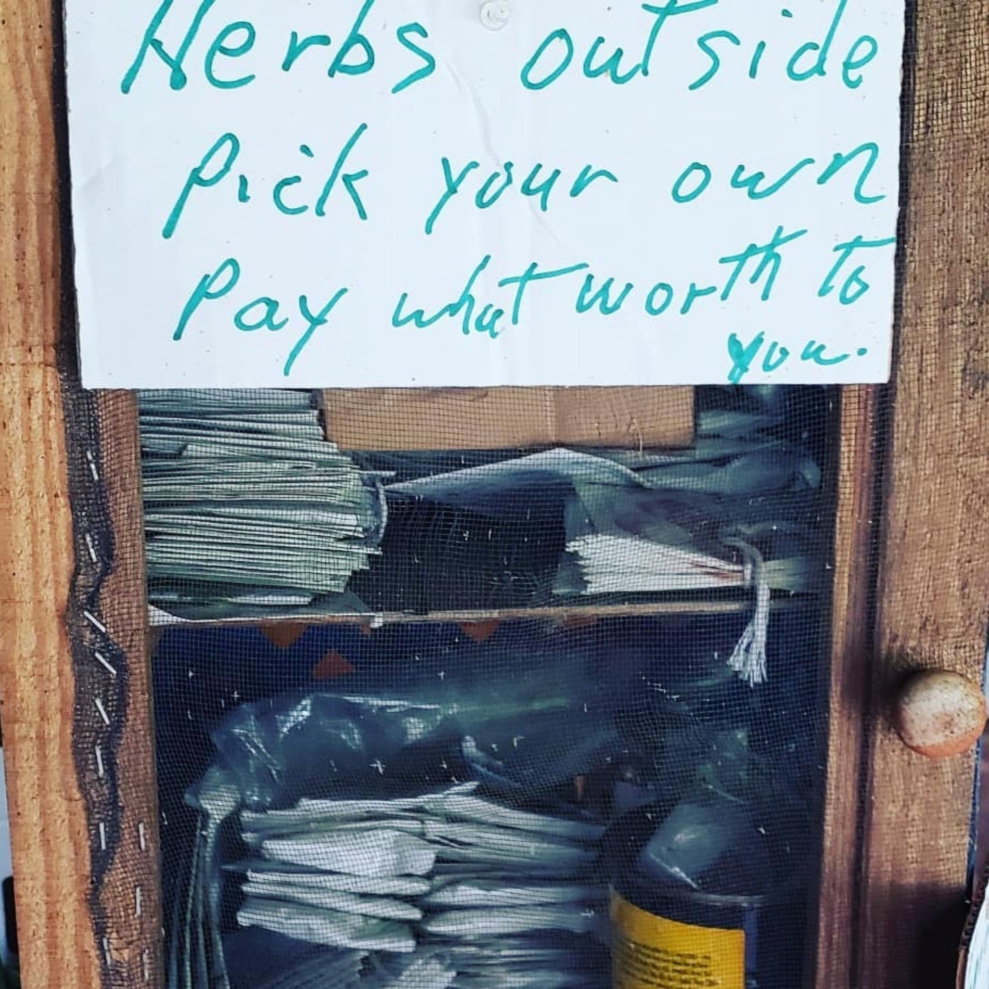 A handwritten sign reads, “Herbs outside. Pick your own. Pay what they’re worth.” Below, Dot Montgillion Memorial Herb Library Membership seed packets and bottles by Smoke Camp Crafts are stored in a rustic wooden cabinet with mesh doors.