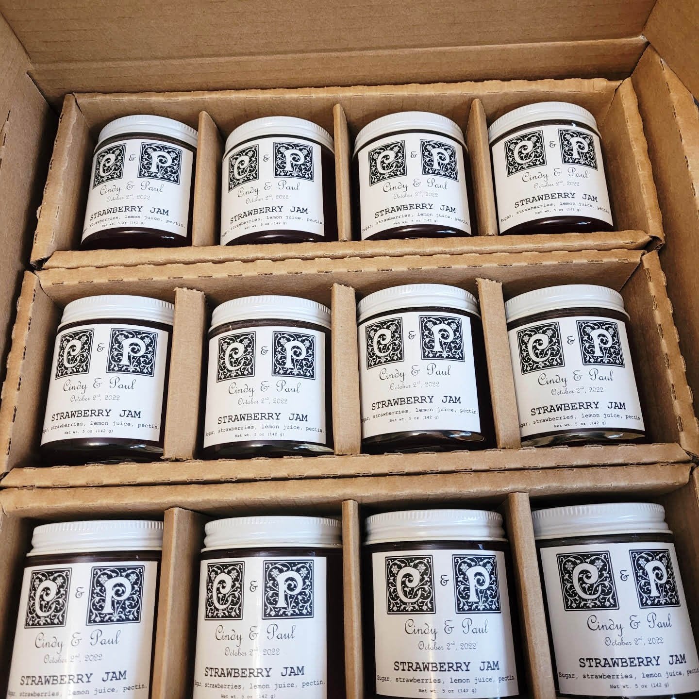 An open box holds twelve jars of Smoke Camp Crafts' Jams and Jellies for Your Special Event, arranged in three rows with paper dividers. Each has a white lid and a custom black-and-white label—ideal as event favors.