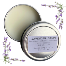 A partially open tin of Smoke Camp Crafts Lavender Salve - 1 oz reveals the cream inside. The label displays ingredients and maker, while sprigs of lavender flowers accent the image’s corners.