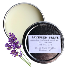 A 1 oz tin of Lavender Salve by Smoke Camp Crafts sits open to reveal creamy balm, with its natural ingredients and origin on the lid. Two fresh lavender sprigs rest beside the tin.