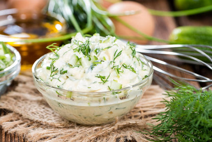 A clear glass bowl of creamy tzatziki sauce, topped with fresh dill and Smoke Camp Crafts Herbal Seasoning Blend Dill Dip Mix, sits on a rustic wooden surface surrounded by herbs, a whisk, cucumbers, and oil.
