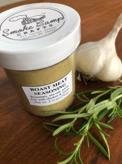 A jar of Smoke Camp Crafts Roast Meat Seasoning, featuring an organic spice blend, rests on a wooden surface next to a whole garlic bulb and a fresh rosemary sprig.
